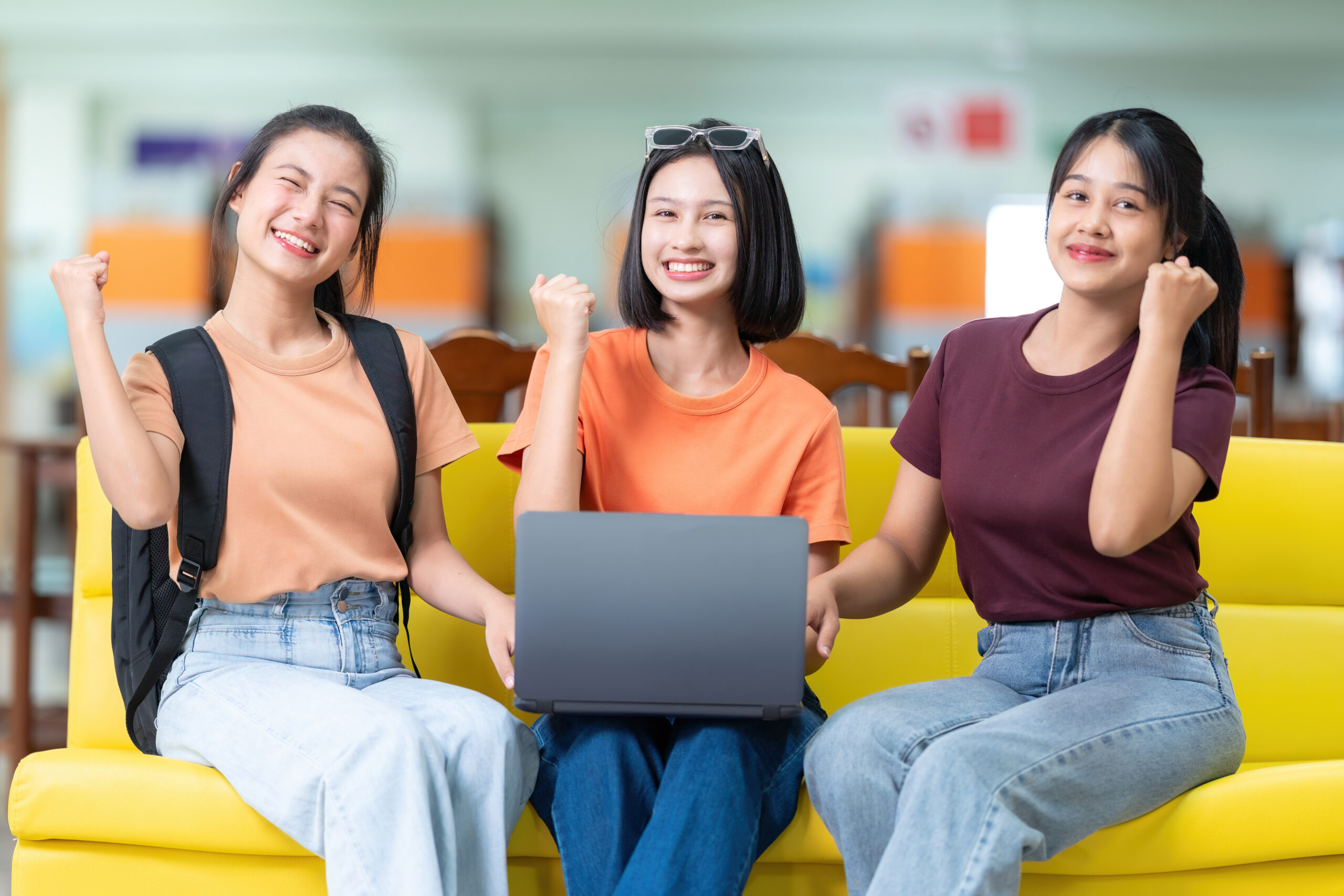 group 3 asian female students excited happy that they passed final exam got excellent grades have laptops university library sitting sofas wearing casual clothes with bags scaled